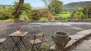A gravel patio with a wooden table and chairs, a stone planter, and a backdrop of trees and hills. - The Chapel - UK45026 (Crickhowell)