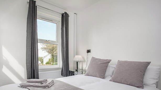 A neatly made bed with a grey blanket and pillows, a window with curtains, a bedside lamp, and a view of the outdoors. - 2 bedroom accommodation in Foxhole, near St. Austell (Foxhole, near St. Austell)