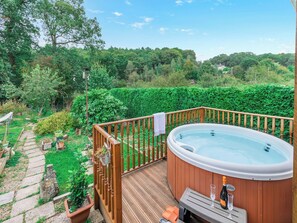 Un jacuzzi en una terraza de madera con barandilla, rodeado de abundante vegetación y un sendero de piedra.