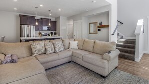 A modern living room with a sectional sofa, a kitchen in the background, and a staircase leading to another level. - New Modern Twin Home, Pet Friendly, Close to Airport, Downtown, Resorts and more (Millcreek)