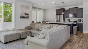 A modern living room with a sectional sofa, a kitchen with dark cabinets, and a large window with a view of trees. - New Modern Twin Home, Pet Friendly, Close to Airport, Downtown, Resorts and more (Millcreek)