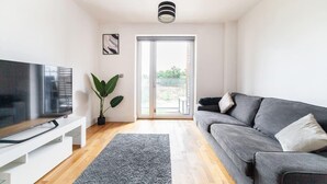 A modern living room with a flat-screen TV, a grey sofa, a large potted plant, and a sliding glass door leading to an outdoor area.
