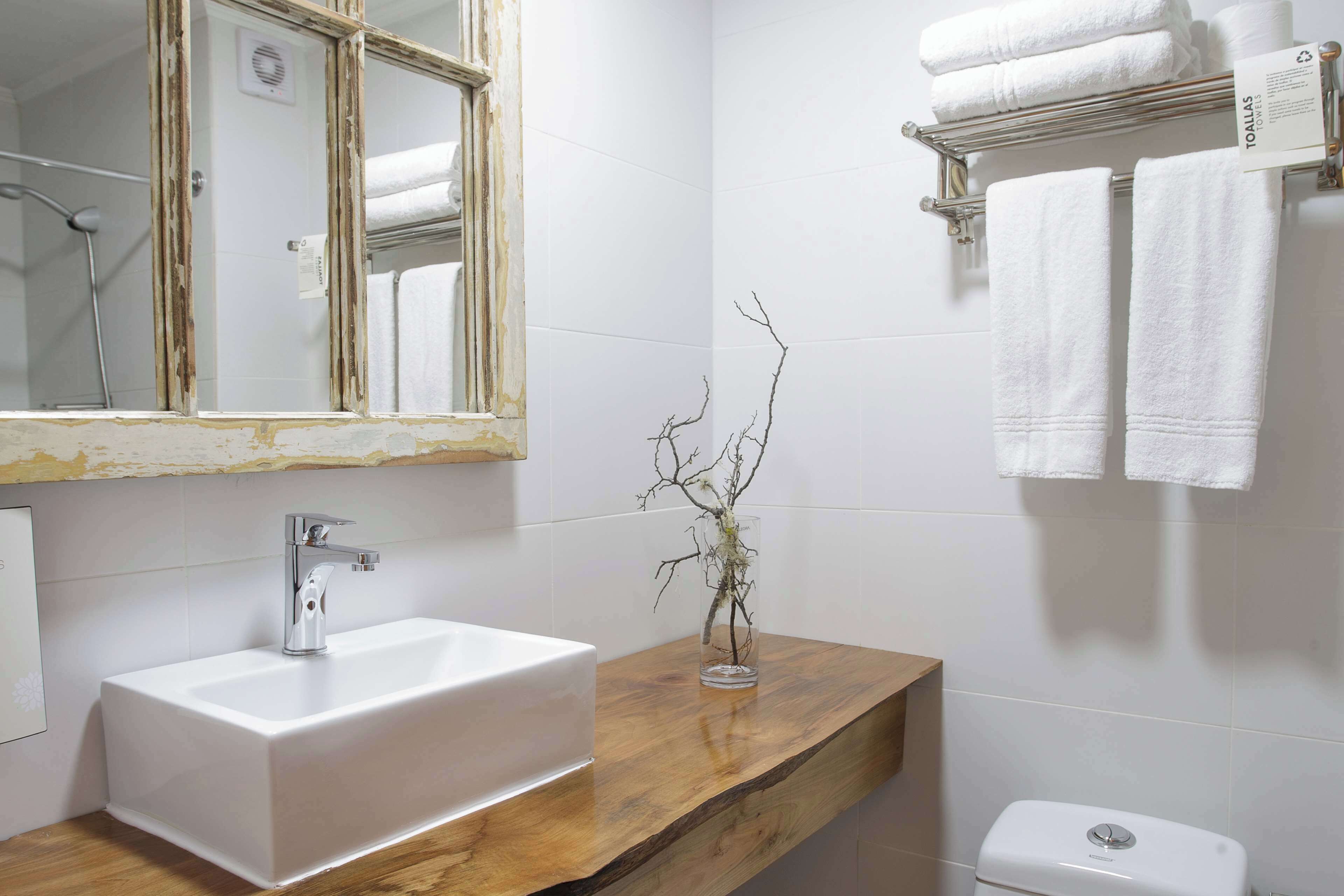 Un baño con un lavamanos blanco, un espejo con marco de madera, un florero de vidrio con ramas y un inodoro.