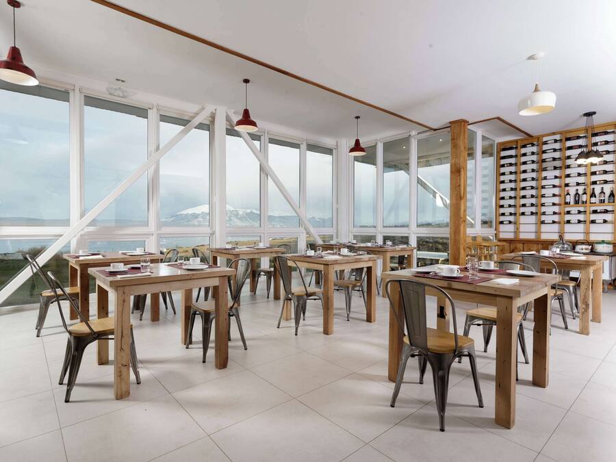 A spacious dining area with multiple tables and chairs, large windows offering a view of a mountainous landscape, and a wall with neatly arranged bottles.