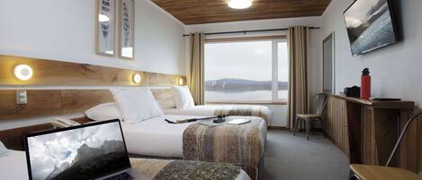 A hotel room with two beds, a desk, a laptop, and a large window with a view of mountains.