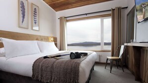 Habitación de hotel con una cama grande, un escritorio con una silla, un televisor y una ventana con vistas a un lago y montañas.