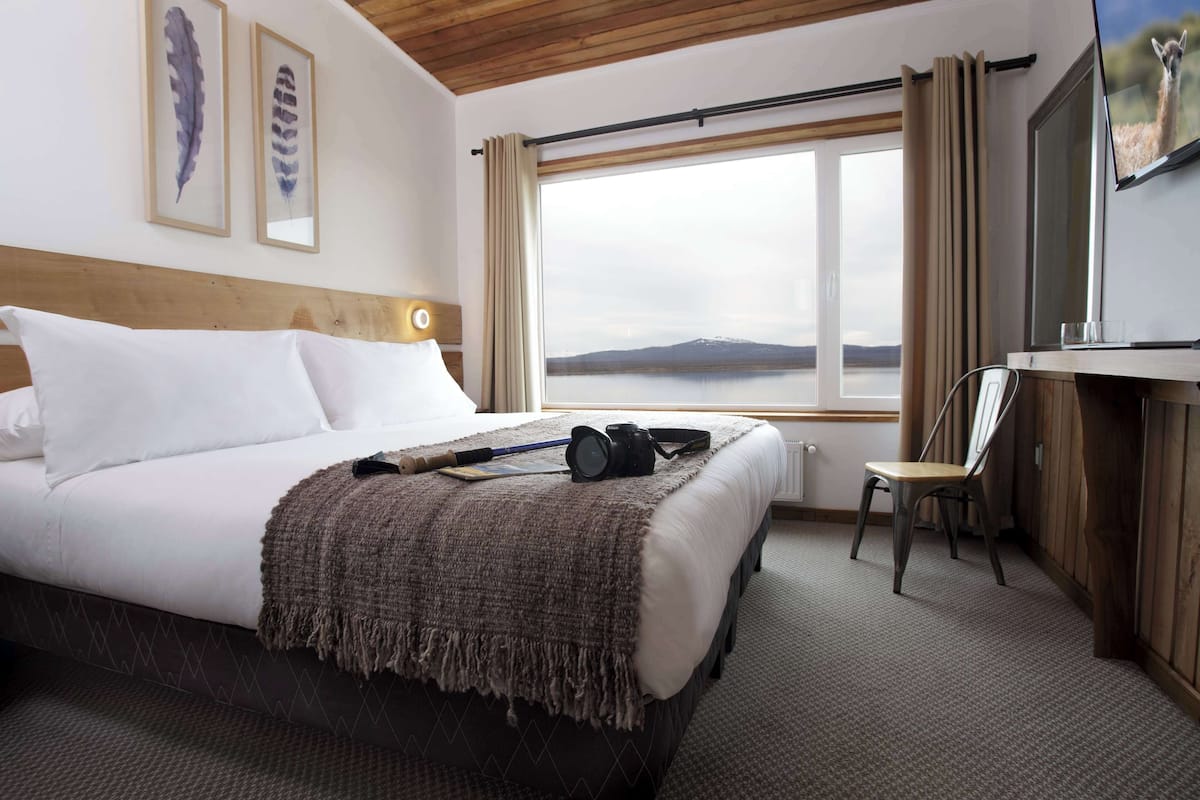 Habitación de hotel con una cama grande, un escritorio con una silla, un televisor y una ventana con vistas a un lago y montañas.