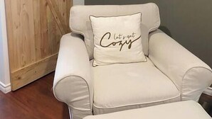 A chair with a cushion that reads "let's get cozy" in a room with a wooden barn door and a plain wall. - The Gathering Place PEC (Prince Edward)