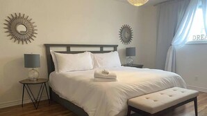 A neatly made bed with white linens, a matching bench, two bedside tables with lamps, and decorative sunburst wall art. - The Gathering Place PEC (Prince Edward)