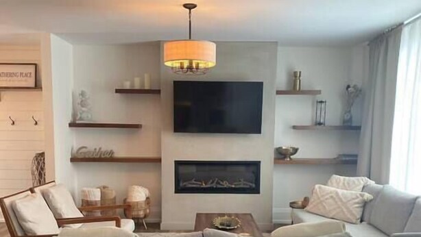 A modern living room with a grey sofa, a flat-screen TV, and a fireplace. There are wooden shelves and a hanging light fixture. - The Gathering Place PEC (Prince Edward)