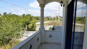 Un balcon donnant sur un paysage verdoyant et une route, un mur blanc avec une porte, et une petite fenêtre.