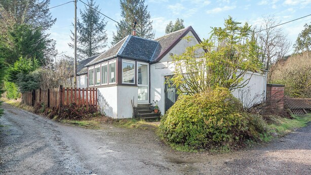 Ein kleines, eingeschossiges Haus mit Satteldach, einer Veranda und einer Auffahrt mit Kies.