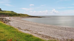 Ein kiesiger Strand mit Rasenflächen und einem entfernten Blick auf das Meer unter blauem Himmel.