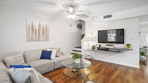 A modern living room with a grey sofa, a glass coffee table, a flat-screen TV mounted on the wall, and a ceiling fan.