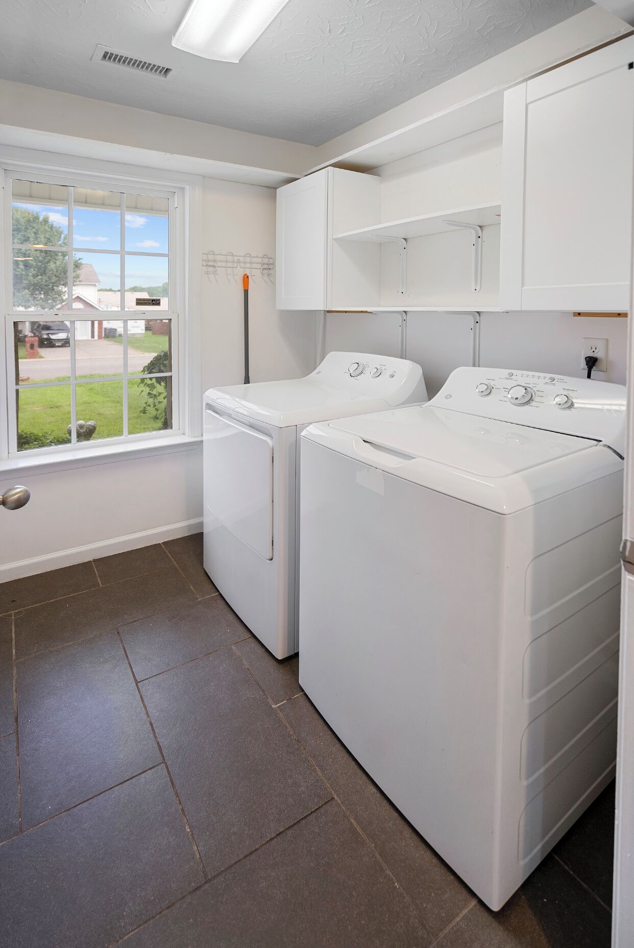 Un cuarto de lavado con dos lavadoras y secadoras blancas, una ventana con vista a una zona residencial, y una puerta con picaporte plateado.
