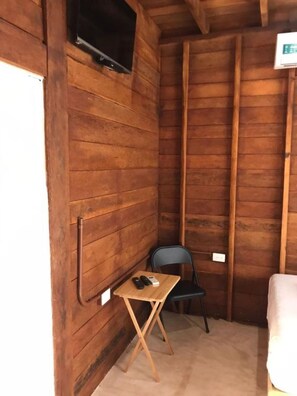 A small wooden room with a single bed, a television mounted on the wall, and a small table with a chair. - Cabanas Chac Mool (Isla Holbox)
