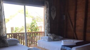 Une chambre avec deux lits en bois, chacun agrémenté de draps et d’oreillers blancs, un plancher en bois et une grande fenêtre donnant sur un paysage verdoyant.