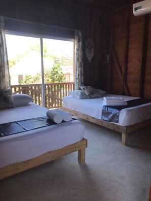A room with two wooden beds, each with white linens and pillows, a wooden floor, and a large window with a view of greenery outside. - Cabanas Chac Mool (Isla Holbox)