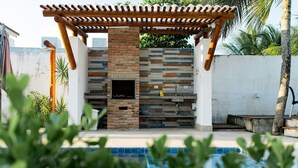 Un espace barbecue extérieur avec une cheminée en briques, une pergola et une piscine en premier plan.