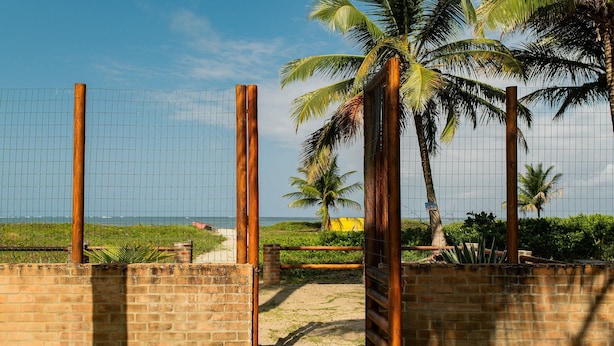 Une entrée fermée par un portail, avec un mur de briques et des poteaux en bois, qui mène à une plage bordée de palmiers et offrant une vue sur un ciel dégagé.
