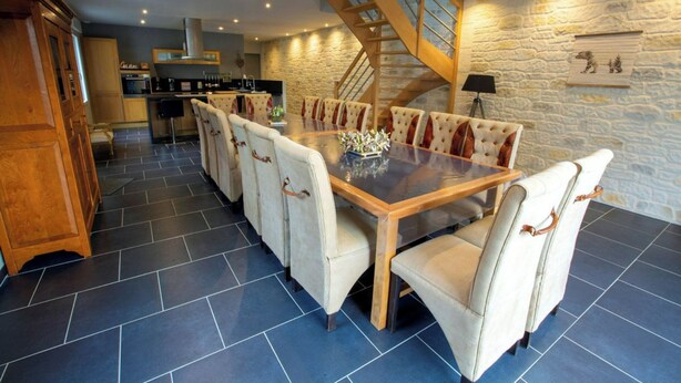 A spacious dining area with a large wooden table and upholstered chairs, a kitchen in the background, and a stone wall with a staircase.