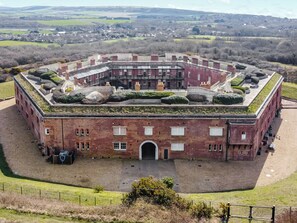 A large, circular brick building with a central courtyard, surrounded by a fence and a grassy area.