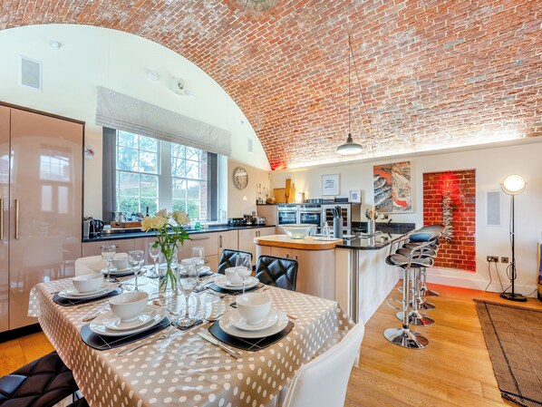 A dining area with a brick ceiling, a kitchen in the background, and a dining table set with white tableware.