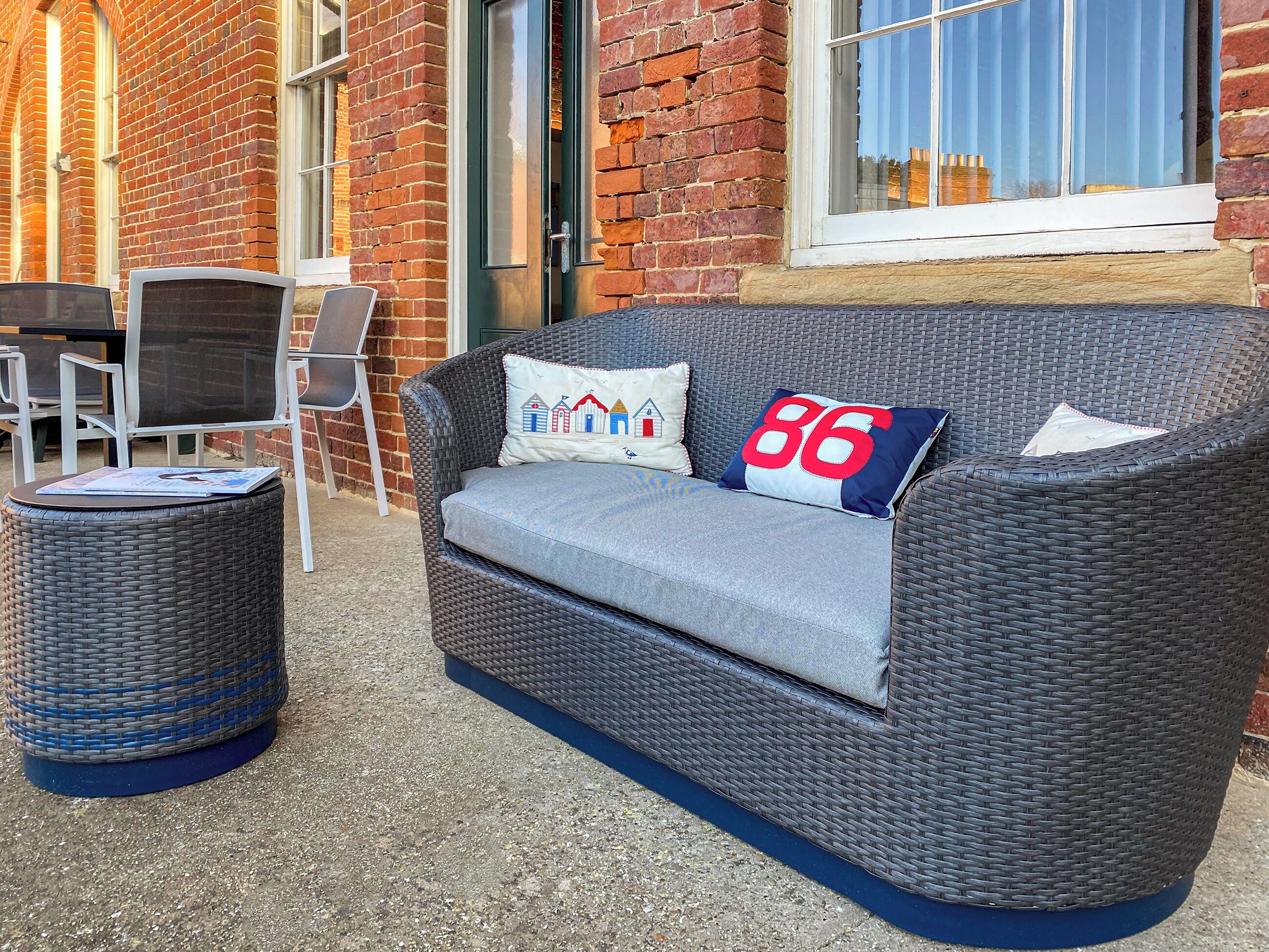 A wicker sofa with cushions, a round wicker table, and a brick wall in the background.