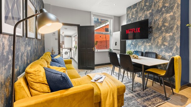 Una sala de estar moderna con un sofá amarillo, una mesa de comedor con sillas, un televisor montado en la pared y vistas al exterior a través de una ventana.