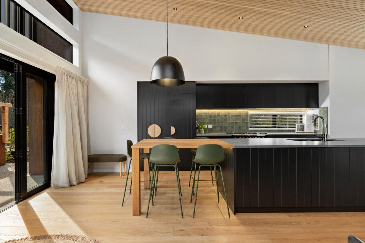 A modern kitchen with a wooden dining table and green chairs, a black pendant light, and a large window with curtains.