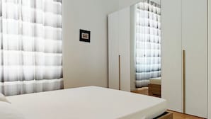 A bedroom with a bed, wooden floor, white walls, a mirror, and a window with curtains. - OXILIA (Milan)
