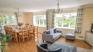 A dining area with a wooden table and chairs, a grey sofa, and a black armchair. There is a window with a view of greenery outside. - Hillside Lodge (Llandrindod Wells)