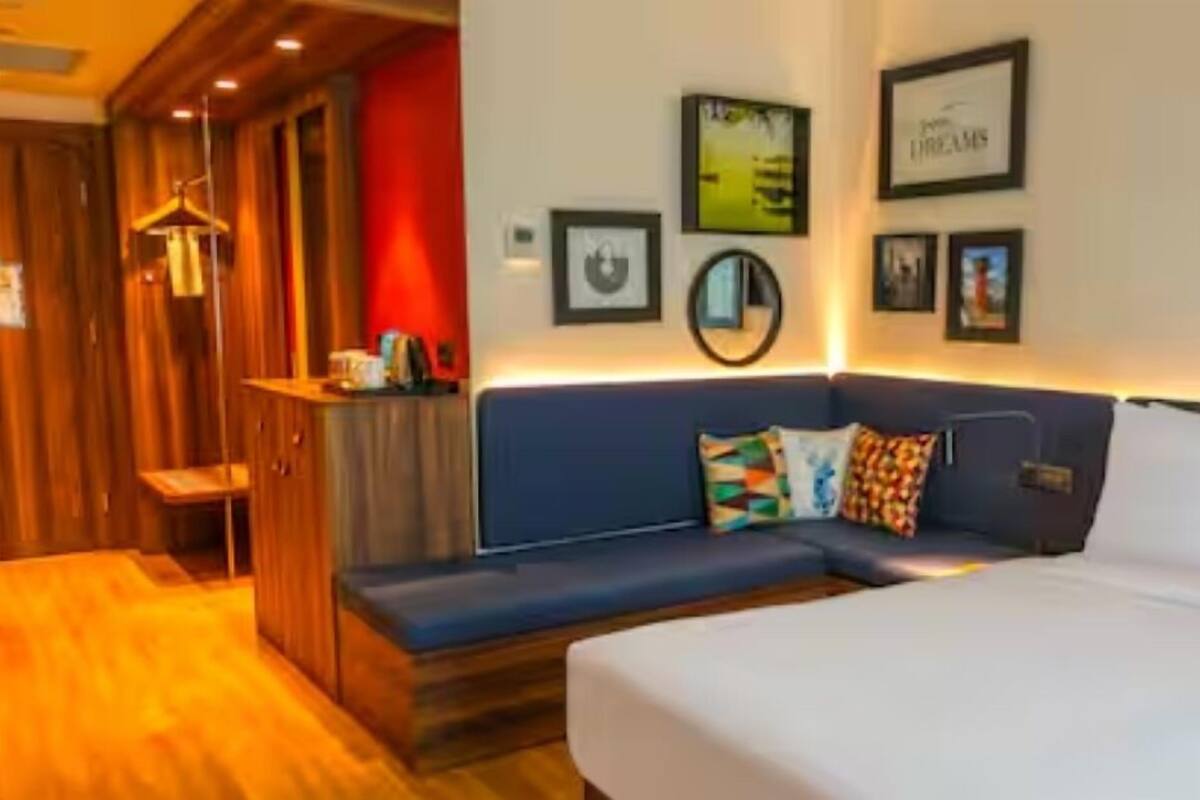 Habitación de hotel con un armario de madera, un sofá azul con cojines coloridos, una cama blanca y obras de arte colgadas en la pared.