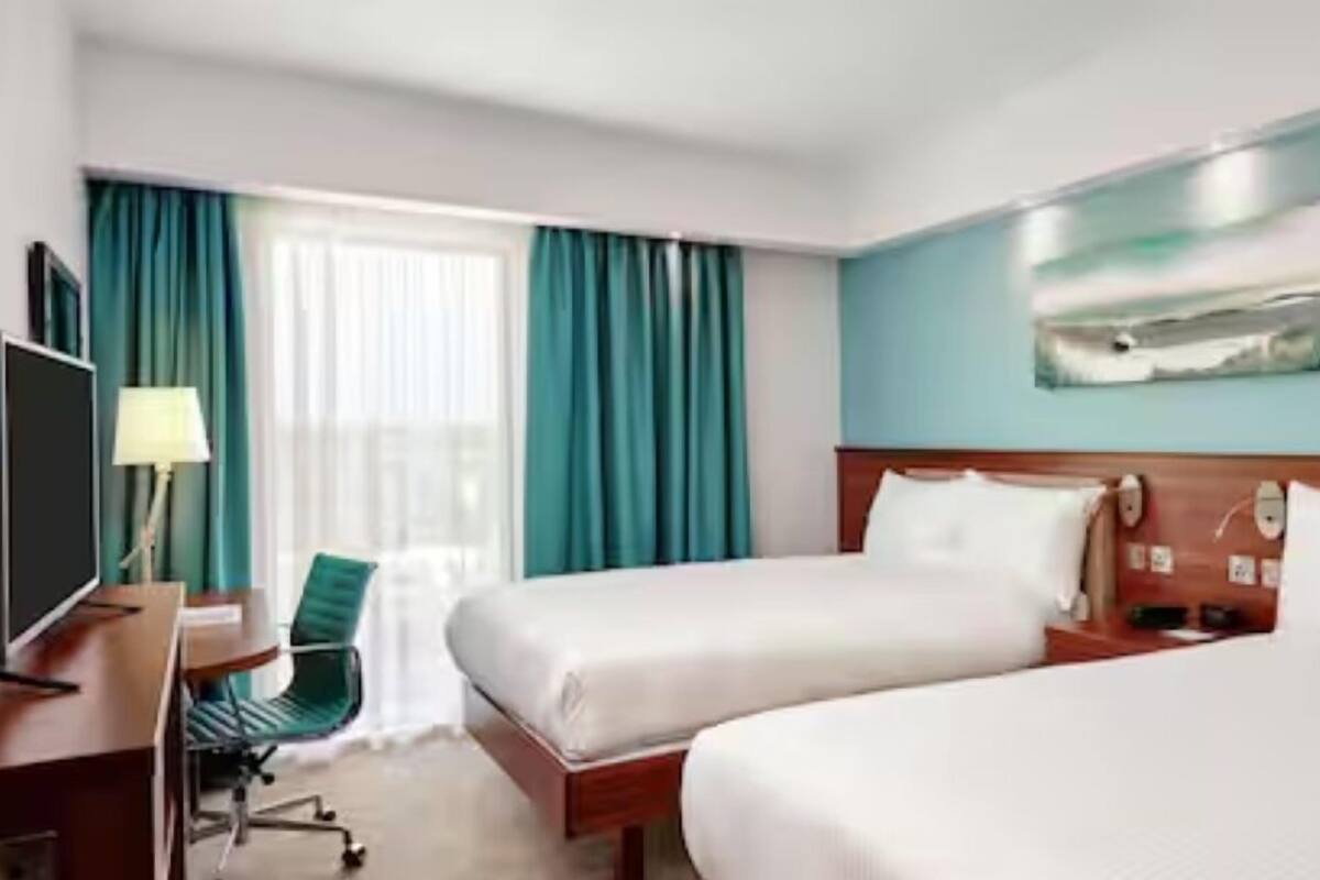 Habitación de hotel con dos camas, un escritorio con silla, un televisor y un cuadro en la pared.