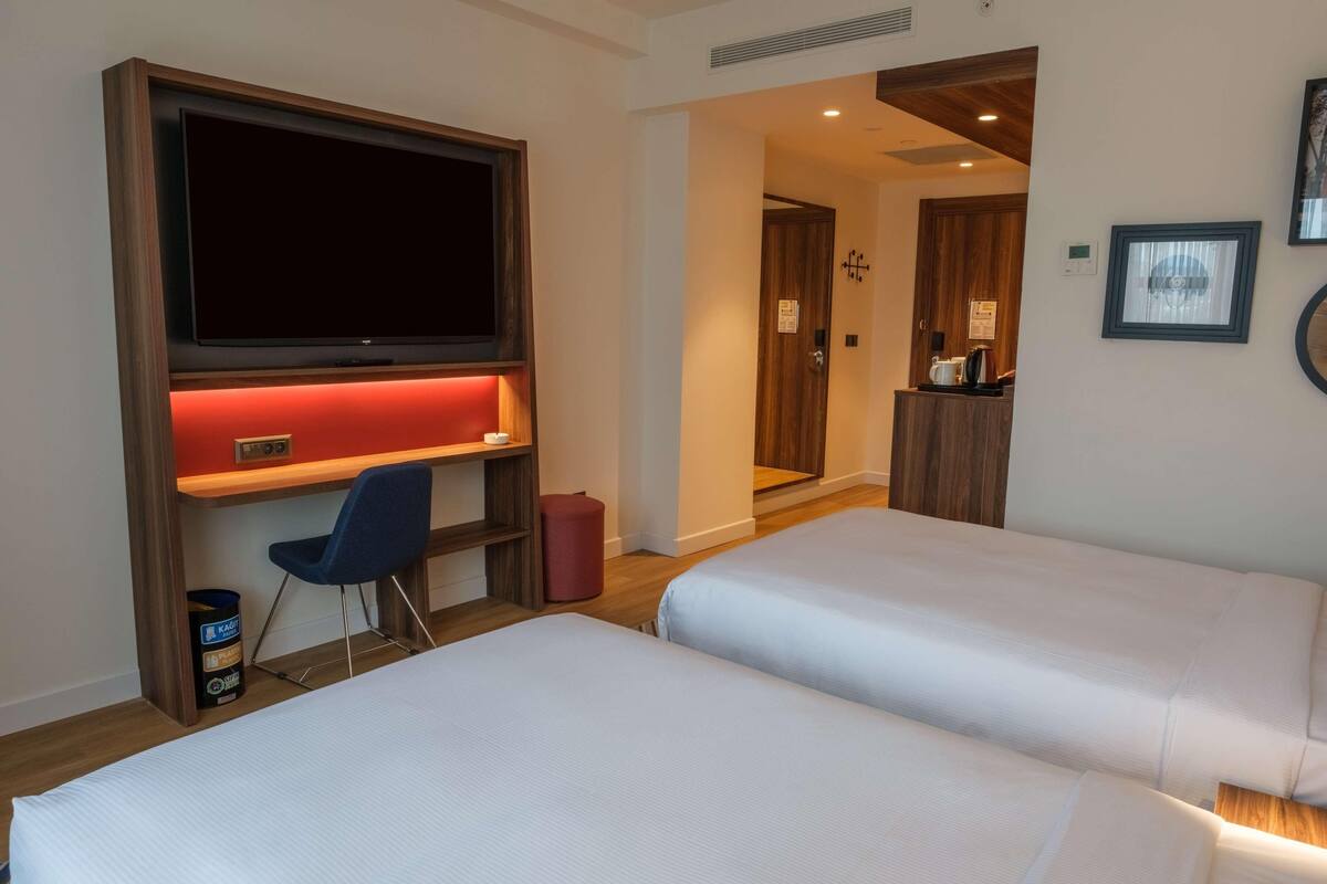 Habitación de hotel con dos camas, un escritorio con silla, televisión y una pequeña cocina.