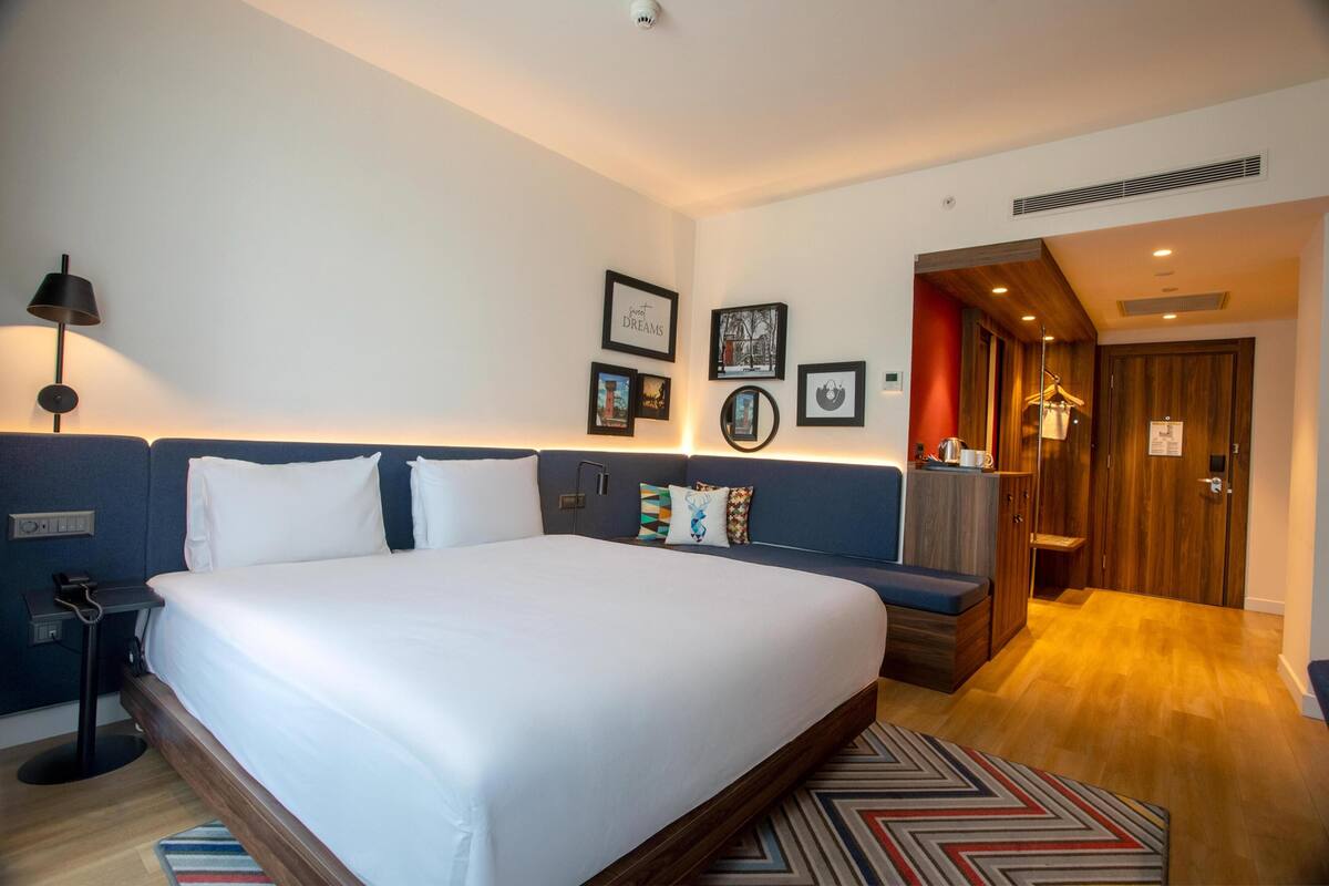 Habitación de hotel con una cama grande, una mesita de noche con lámpara, un armario de madera y una pequeña zona de estar con un banco.
