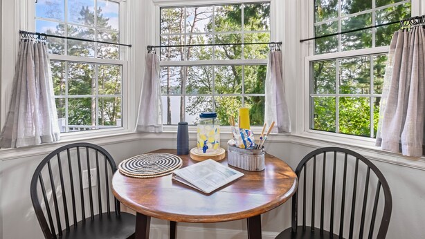 A dining area with a round table, two chairs, a window with curtains, and a view of trees. - Lake Front Cottage with Amazing Firepit, Private Dock, Sleeps 6 (Chippewa Falls)