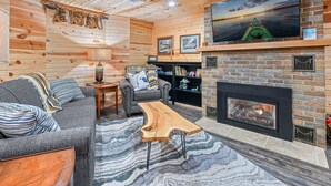 A cozy living room with a fireplace, wooden walls, a sofa, and a TV mounted above the fireplace. - Lake Front Cottage with Amazing Firepit, Private Dock, Sleeps 6 (Chippewa Falls)