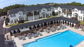 A swimming pool with lounge chairs and umbrellas, surrounded by multi-story buildings with balconies. - Enjoy The Ocean Breeze just Steps from the Beach! (Corolla)