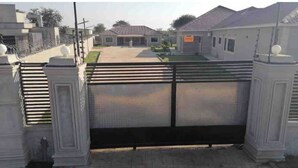 A gated entrance with a metal gate, a paved driveway, and a building with a tiled roof in the background. - Grace’s Cozy Place Unit A in Makeni, Lusaka (Lusaka)