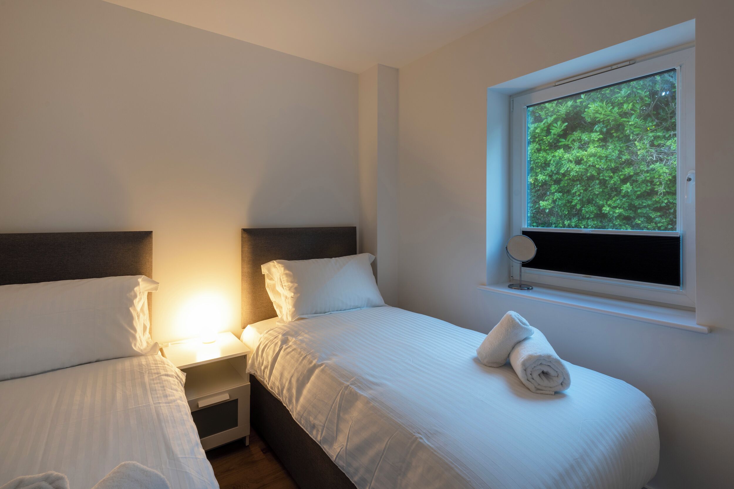 Un dormitorio con dos camas, una mesita de noche, una ventana con vistas a la vegetación y una lámpara fijada a la pared.