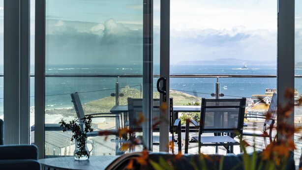 A room with a large window overlooking a coastal view, including a body of water and distant land. - Treen (St Ives)