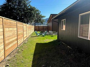 Un patio trasero cercado con una terraza de madera, un pequeño patio con sillas y una mesa, y un edificio con una ventana.