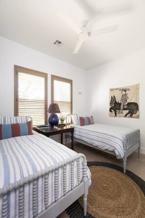 A bedroom with two single beds, a ceiling fan, a window with blinds, a wall-mounted lamp, and a tapestry. - NEW urban cottage, The Vine & Round Top! (Austin County)