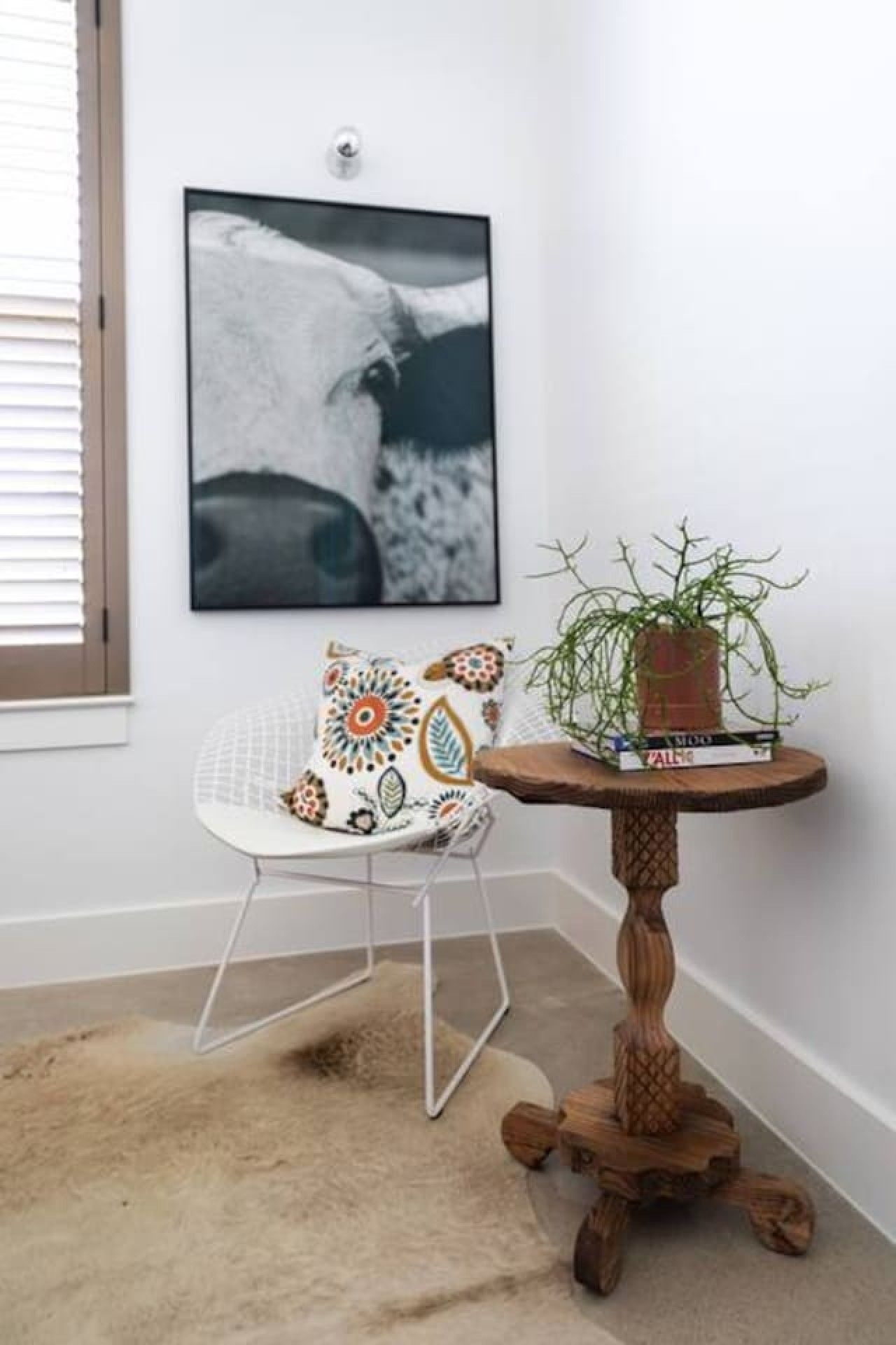 Une pièce d’angle avec une chaise blanche, une table ronde en bois ornée d’une plante, et une photographie en noir et blanc accrochée au mur.