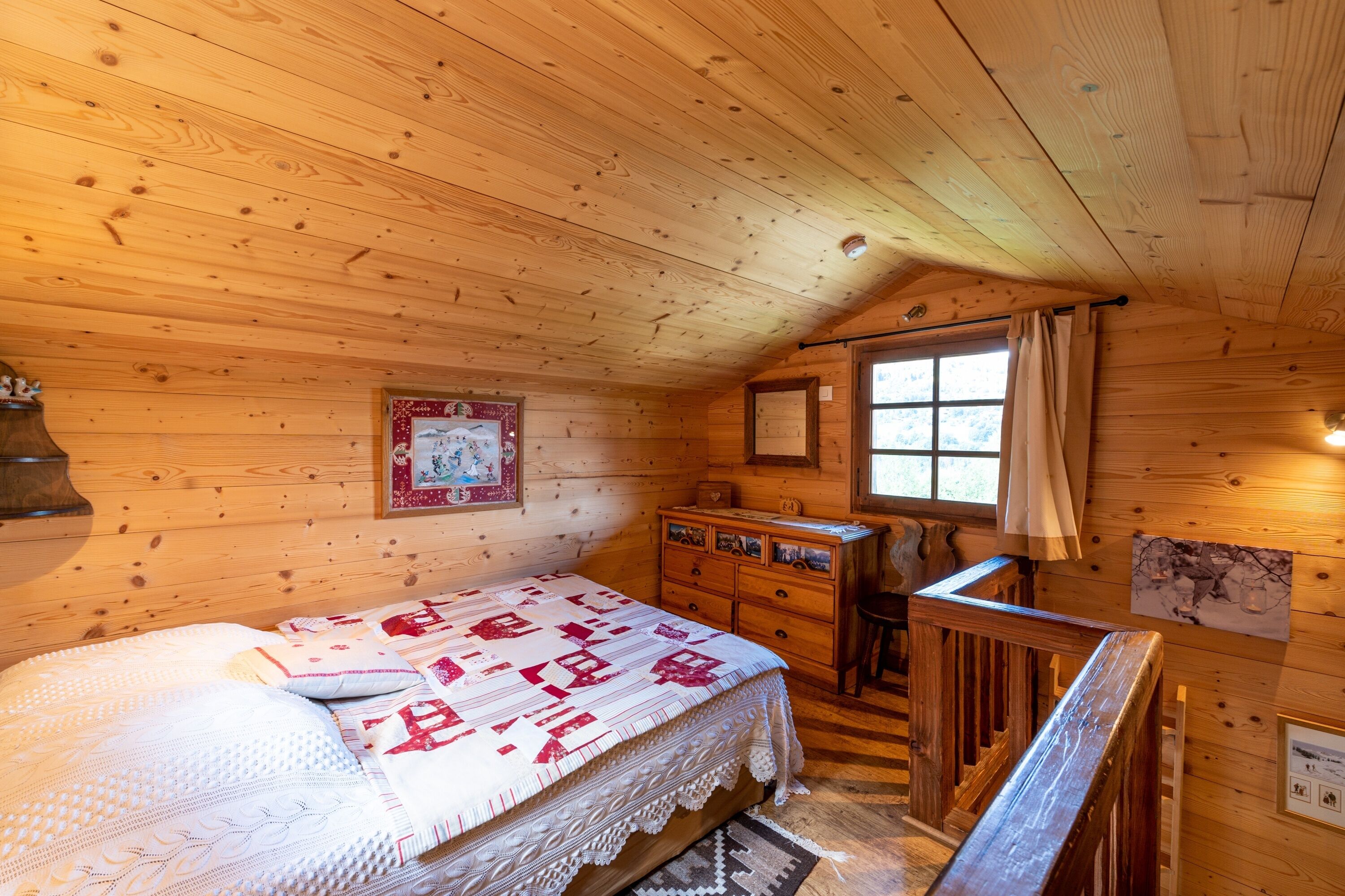 Een slaapkamer in een houten hutje, met een bed, een dressoir en een raam met gordijnen.