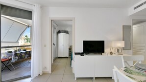 Una sala de estar moderna con televisor, muebles blancos, un balcón con mesa y vistas al exterior.