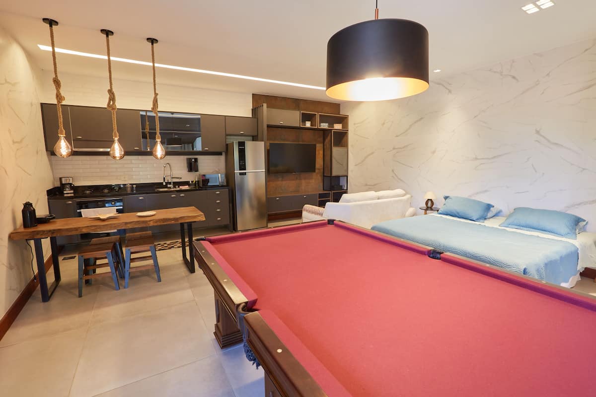 A modern hotel room with a pool table, a dining area, a kitchen, and a large bed with blue bedding.