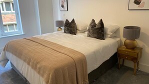 A bedroom with a large bed, two pillows, a bedside lamp, and a small wooden nightstand. - Apartment 1 (York)