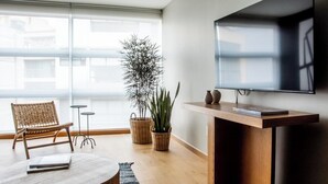 Un salon moderne doté d’une grande fenêtre, d’un téléviseur à écran plat, d’une table basse en bois, d’un fauteuil en osier et de deux plantes en pot.